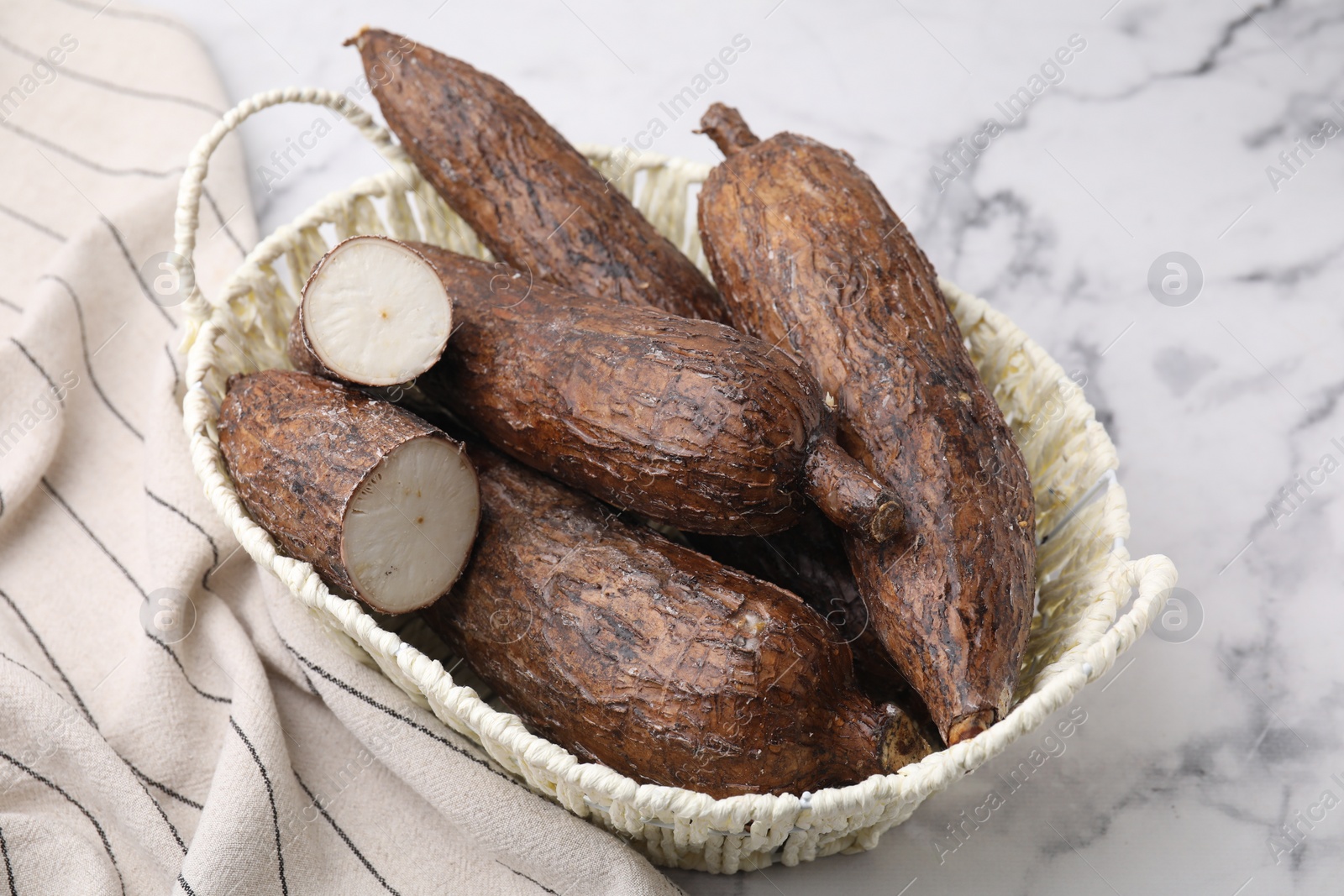 Photo of Whole and cut cassava roots in wicker basket on white marble table