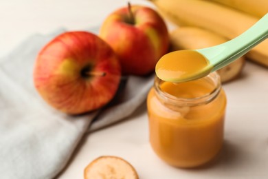 Photo of Spoon with healthy baby food over glass jar on white wooden table, closeup