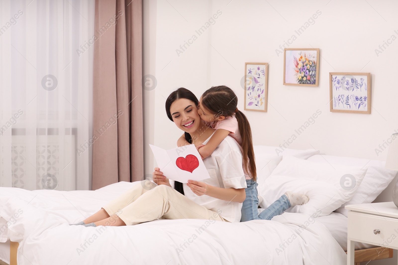 Photo of Happy woman with her daughter and handmade greeting card on bed at home. Mother's day celebration