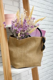 Photo of Stylish beach bag with beautiful bouquet of wildflowers, sunglasses and magazines on shelf indoors
