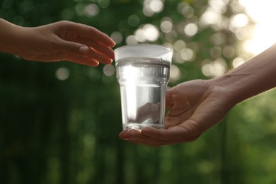 Photo of Man giving woman glass of fresh water in forest on sunny day, closeup