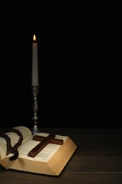 Church candle, Bible, rosary beads and cross on wooden table against black background, space for text