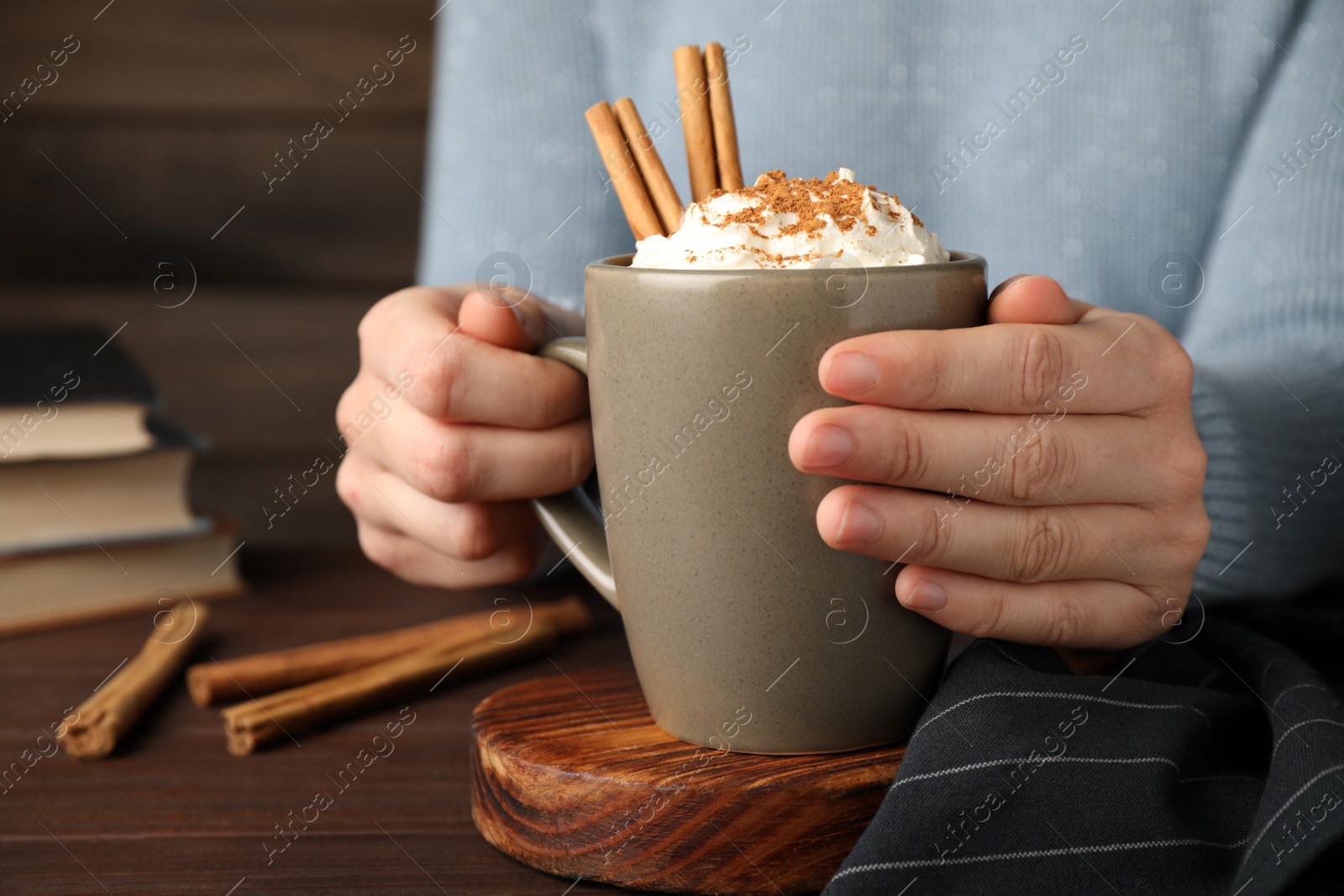 Photo of Woman holding cup of hot drink with aromatic cinnamon and whipped cream at wooden table, closeup