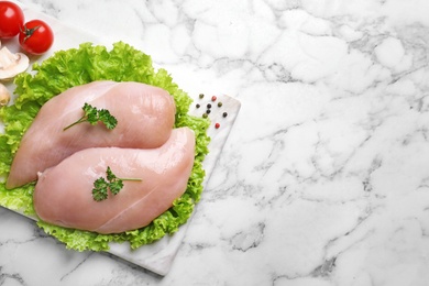 Photo of Board with raw chicken breasts and ingredients on marble background, top view. Space for text