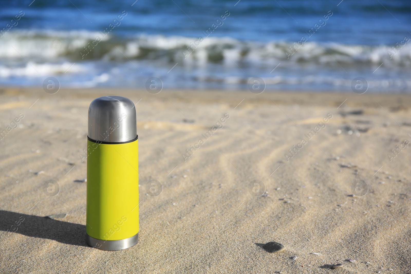 Photo of Yellow thermos with hot drink on sand near sea, space for text