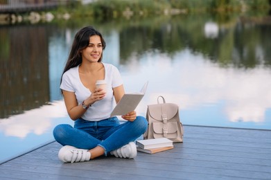 Young woman with cup of coffee reading book on pier near lake