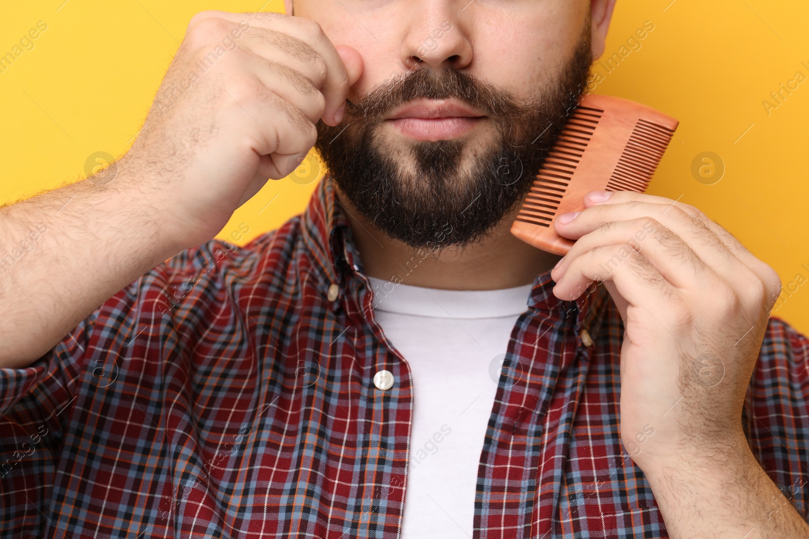 Photo of Handsome young man combing beard on yellow background, closeup