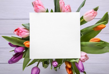 Beautiful bouquet of tulips with blank card on white wooden background, top view. Birthday celebration