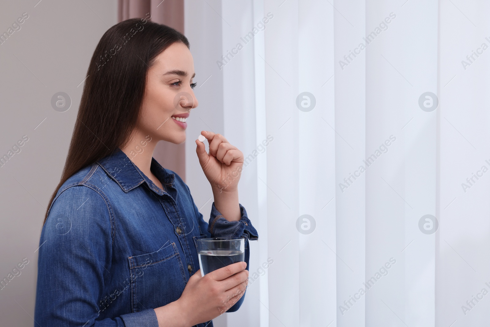 Photo of Beautiful young woman with glass of water and pill indoors, space for text