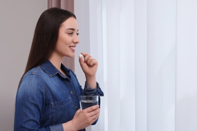 Beautiful young woman with glass of water and pill indoors, space for text