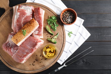 Pieces of raw pork belly, chili pepper, peppercorns, oil and parsley on black wooden table, top view