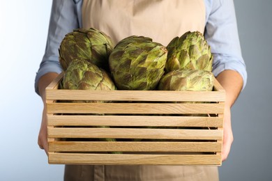 Photo of Woman holding wooden crate with fresh raw artichokes on grey background, closeup