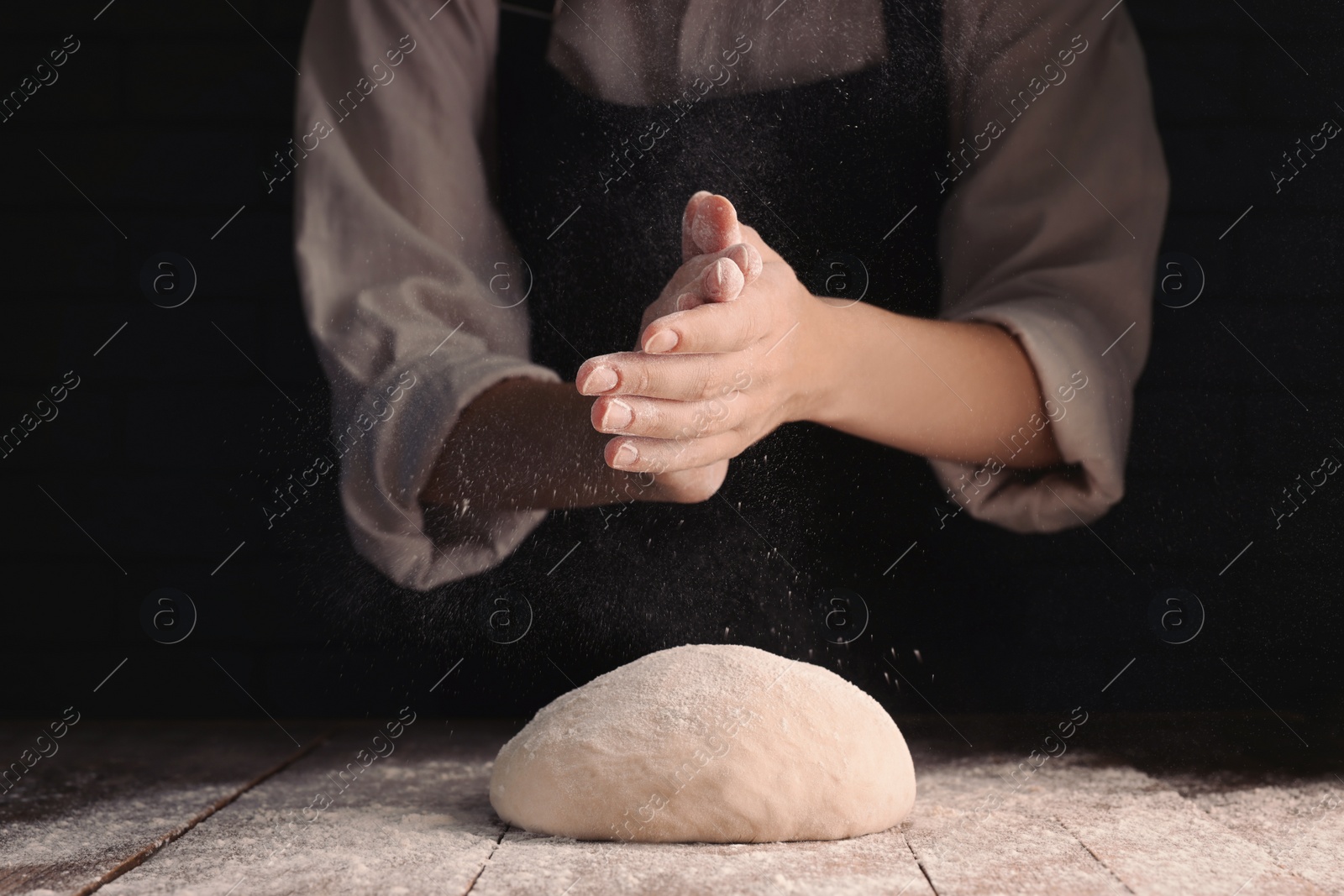 Photo of Woman sprinkling flour over dough at wooden table on dark background, closeup