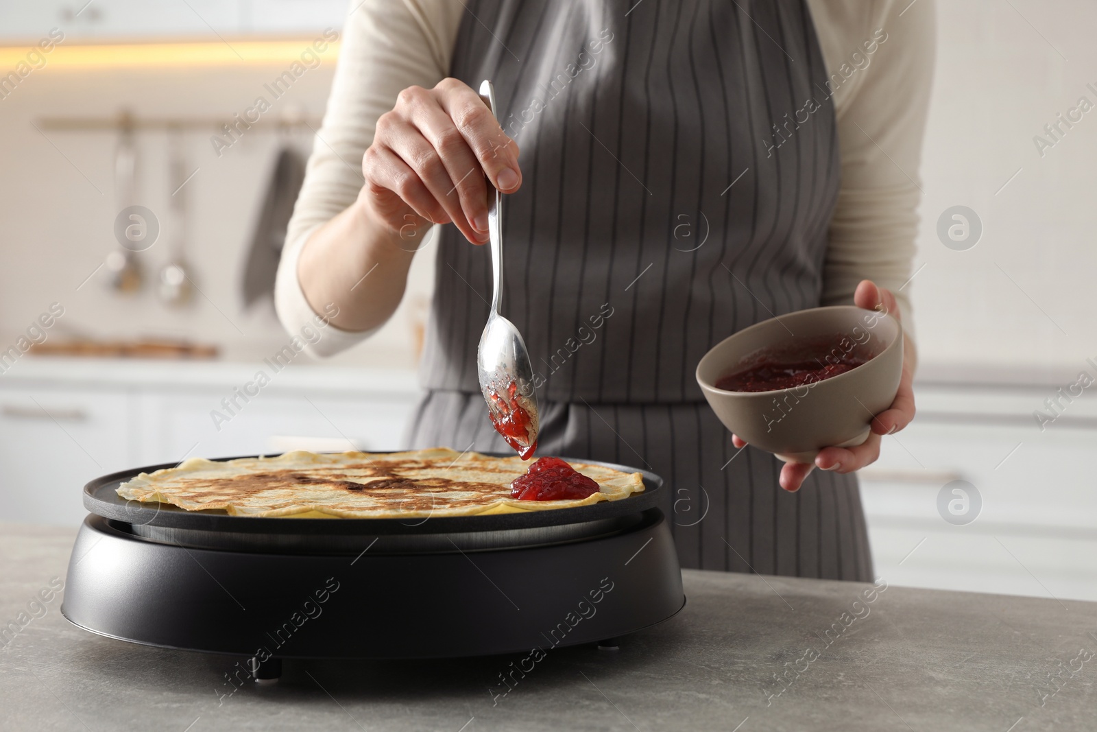 Photo of Woman cooking delicious crepe with jam on electric pancake maker in kitchen, closeup