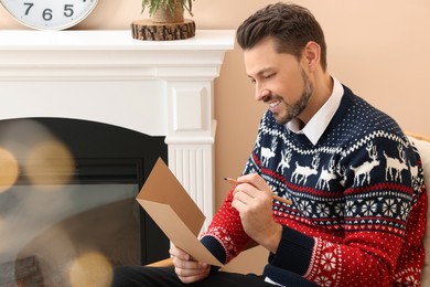Photo of Happy man writing wishes in Christmas greeting card in living room