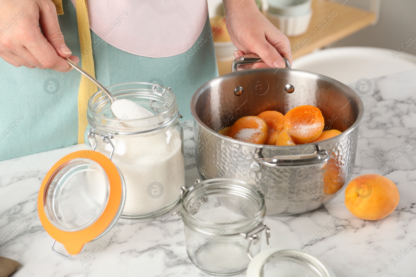 Photo of Woman adding sugar into pot with apricots at table in kitchen, closeup. Making delicious jam