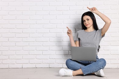 Photo of Student with laptop sitting on floor and pointing at something near white brick wall. Space for text