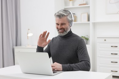 Photo of Man in headphones waving hello during video chat via laptop at home