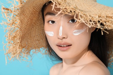 Beautiful young woman in straw hat with sun protection cream on her face against light blue background, closeup