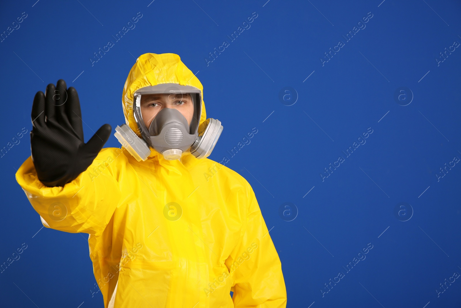 Photo of Man in chemical protective suit making stop gesture on blue background, space for text. Virus research