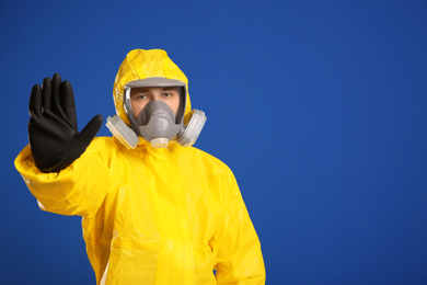 Photo of Man in chemical protective suit making stop gesture on blue background, space for text. Virus research