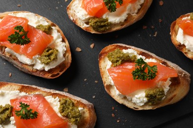 Photo of Delicious bruschettas with cream cheese, salmon and pesto sauce on black table, flat lay