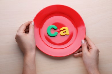 Photo of Woman holding plate with paper symbol Ca (Calcium) at wooden table, top view