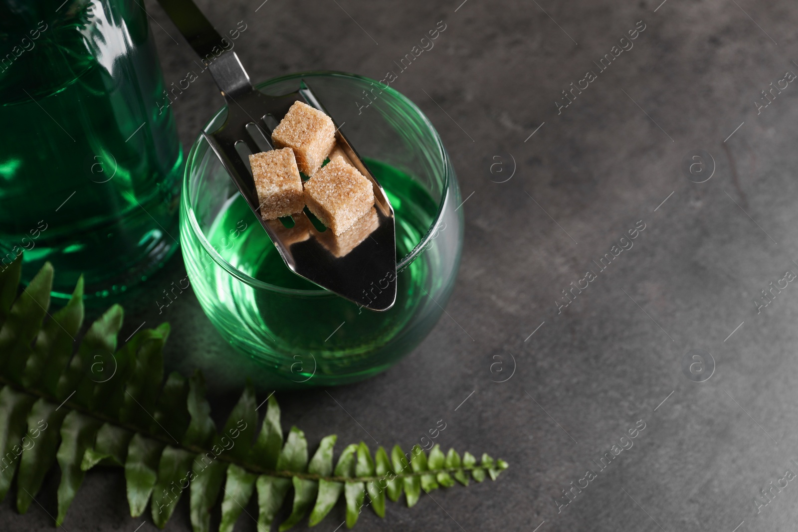 Photo of Absinthe in glass, spoon, brown sugar cubes, and fern leaf on gray textured table, above view with space for text. Alcoholic drink