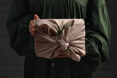 Photo of Furoshiki technique. Woman holding gift wrapped in fabric with thuja branches near dark brick wall, closeup