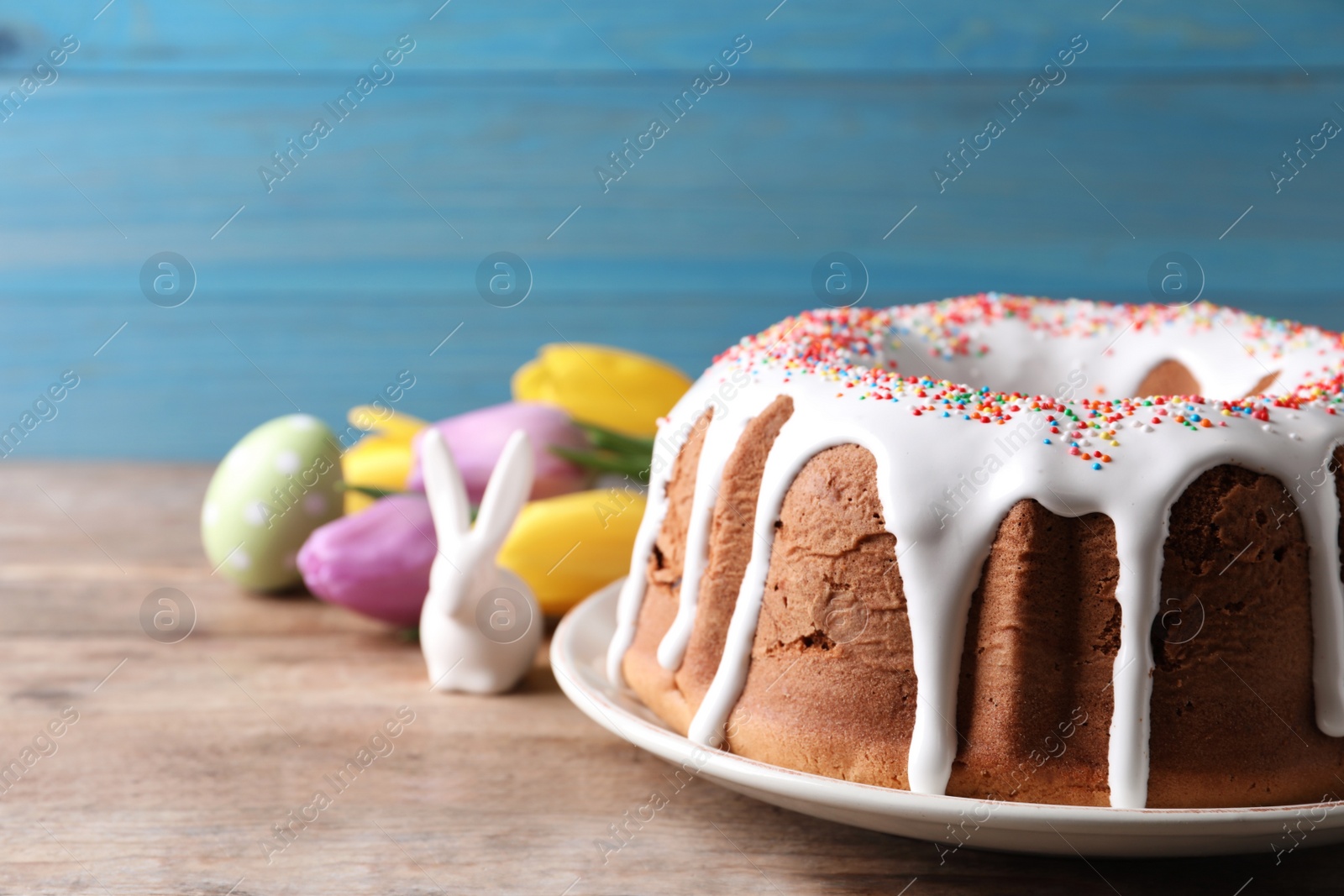 Photo of Glazed Easter cake with sprinkles, decorative bunny, painted egg and tulips on wooden table, closeup. Space for text