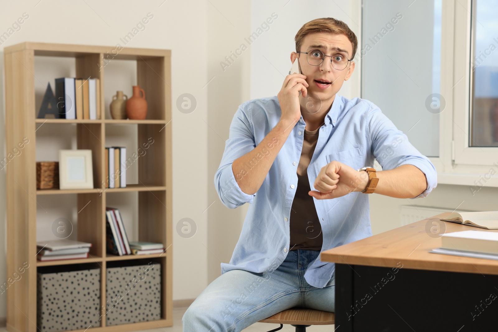 Photo of Emotional young man checking time while talking on phone in office. Being late