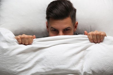 Handsome man covering his face with blanket while lying in bed at home