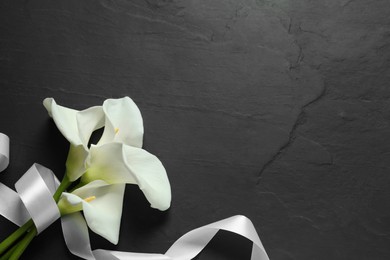 Photo of Beautiful calla lily flowers and white ribbon on black table, flat lay with space for text. Funeral symbols