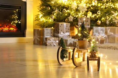 Wooden decorative sleighs in front of many gift boxes, festive lights and Christmas tree on floor. Space for text