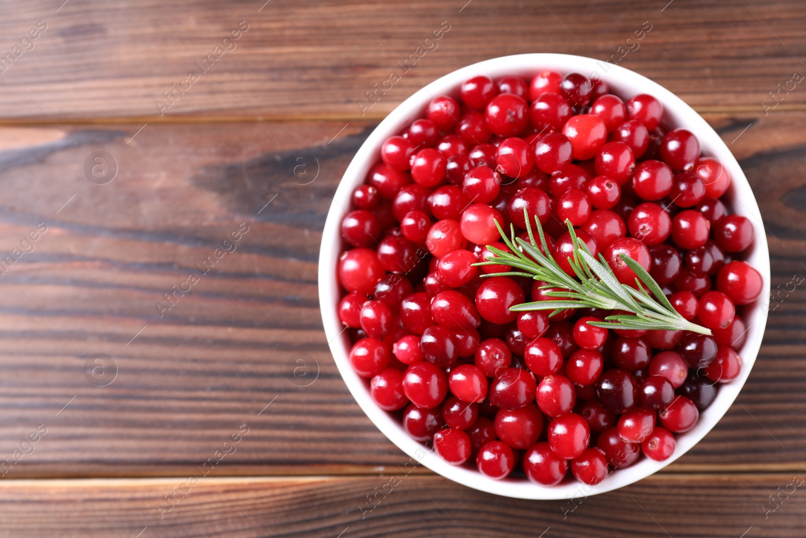 Photo of Fresh ripe cranberries and rosemary in bowl on wooden table, top view. Space for text