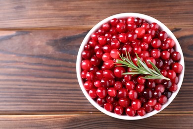 Fresh ripe cranberries and rosemary in bowl on wooden table, top view. Space for text