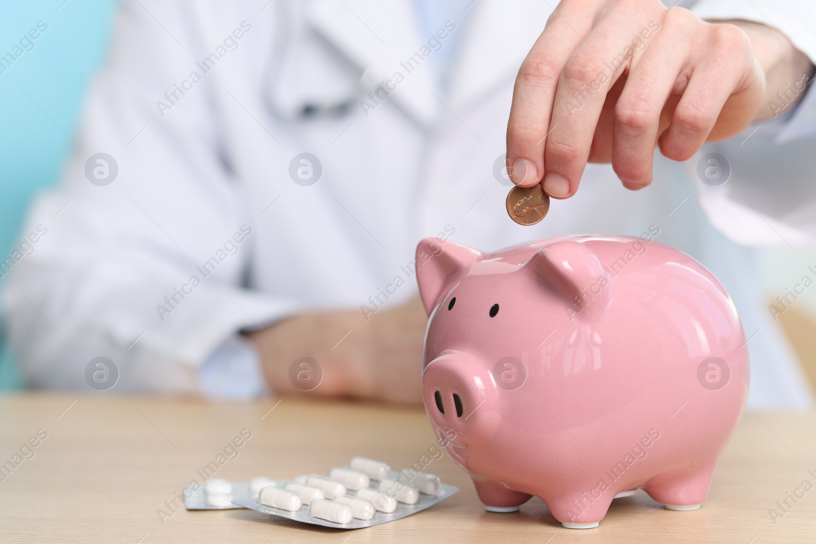 Photo of Doctor putting coin into piggy bank at wooden table, closeup