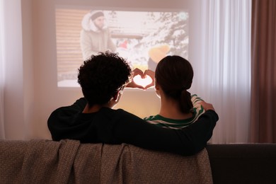 Photo of Couple watching romantic Christmas movie via video projector and making heart with hands at home, back view