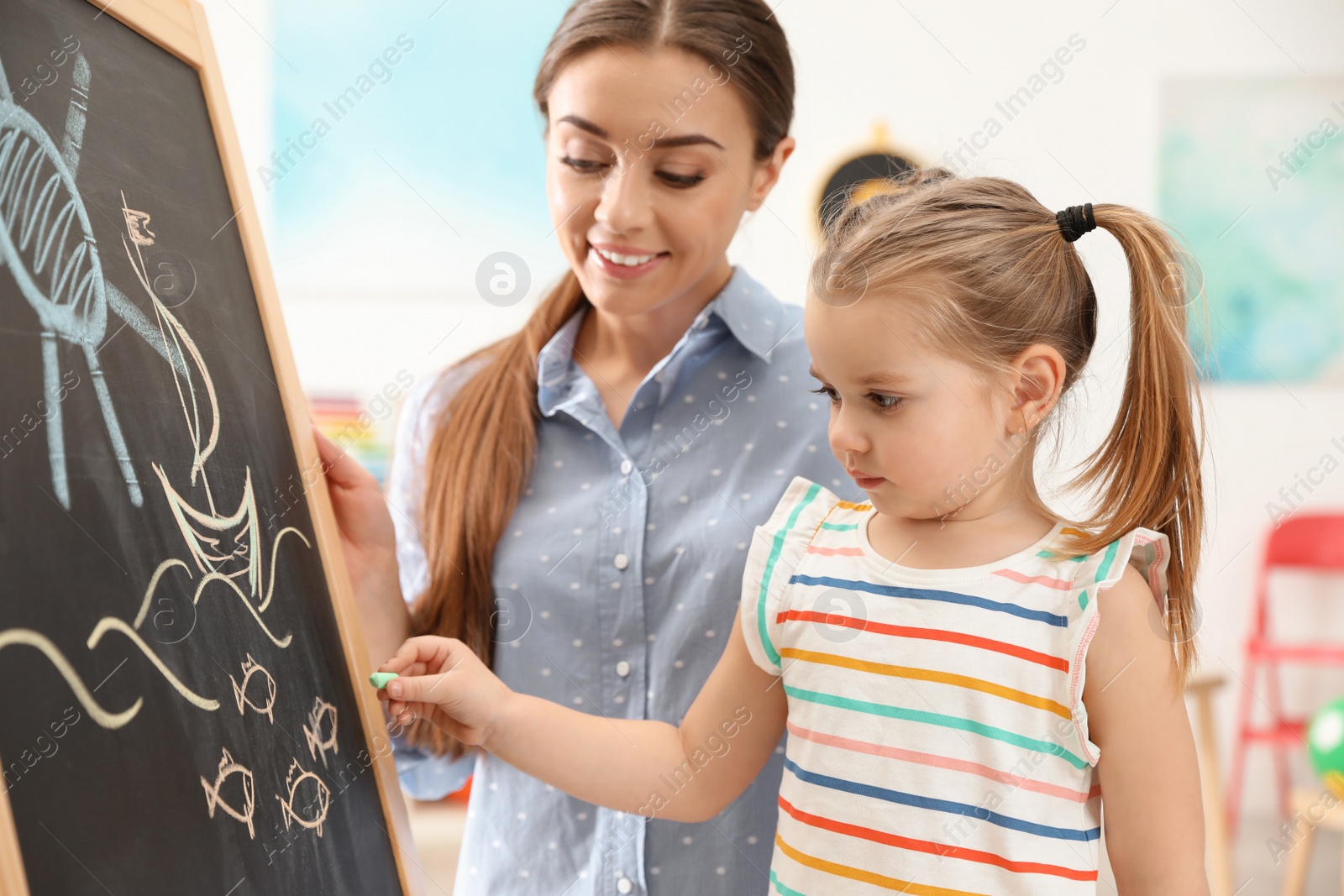 Photo of Kindergarten teacher and little child near chalkboard indoors. Learning and playing