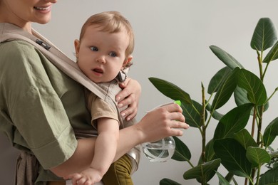 Mother spraying houseplant with water while holding her child in sling (baby carrier) indoors