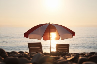 Photo of Empty sunbeds and umbrella on stone beach near sea at sunset