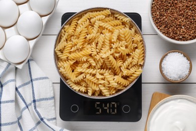 Electronic scales with uncooked pasta and different products on white wooden table, flat lay