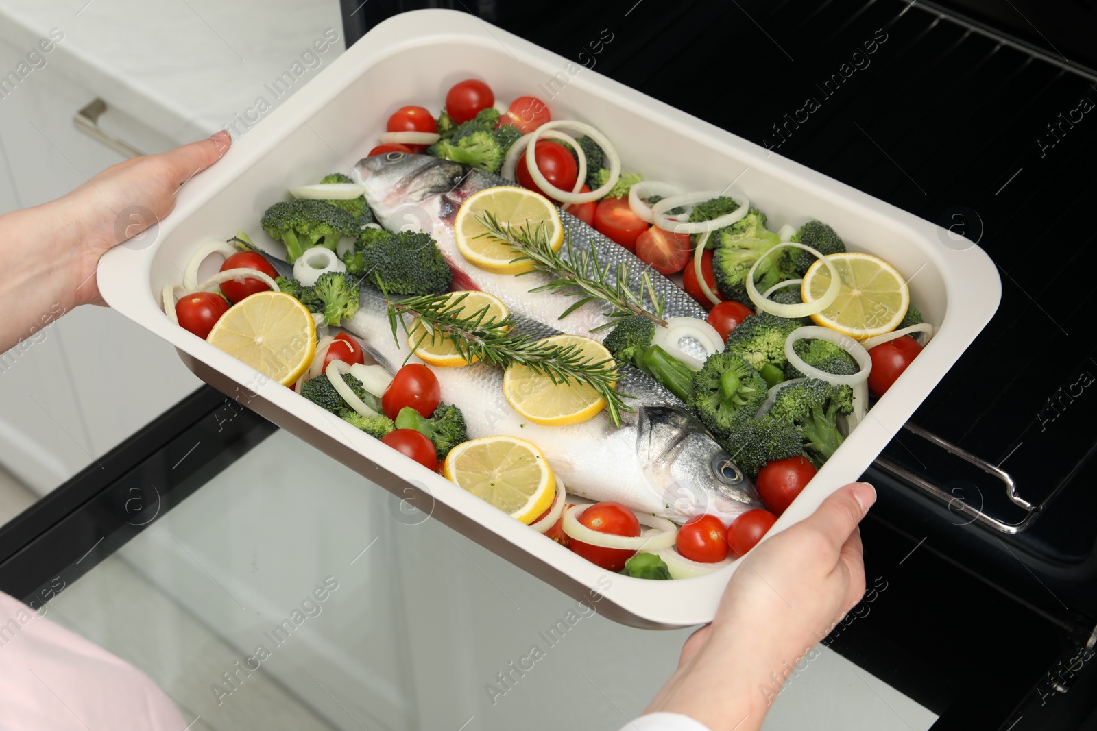 Photo of Woman putting baking dish with raw fish and vegetables into oven in kitchen, closeup