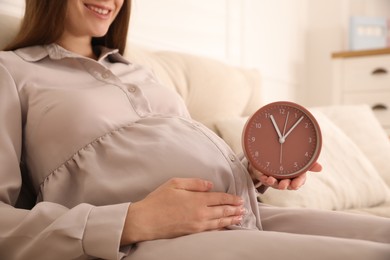 Photo of Young pregnant woman holding clock near her belly at home, closeup. Time to give birth