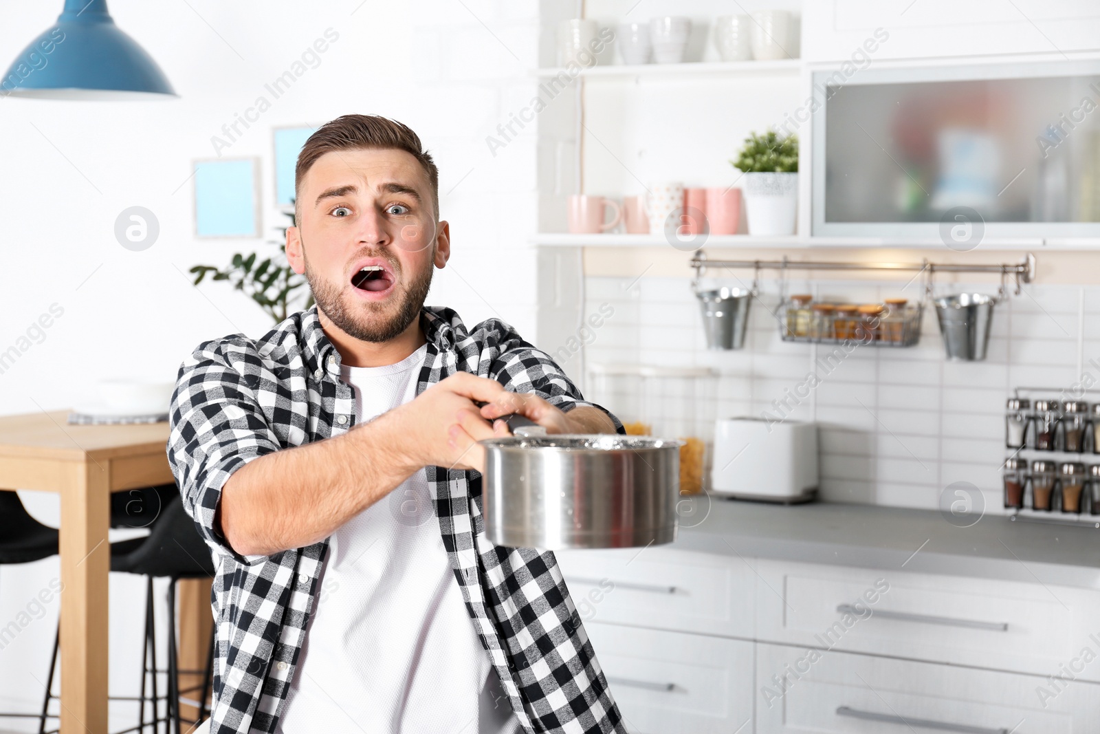 Photo of Emotional young man holding saucepan under water leakage from ceiling in kitchen, space for text. Plumber service