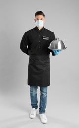 Photo of Waiter in medical face mask holding tray with lid on light grey background