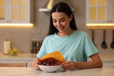 Happy woman packing bowl of fresh grapes into beeswax food wrap at table in kitchen