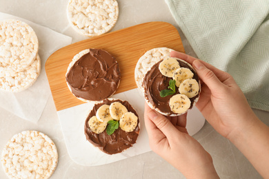 Photo of Woman holding puffed rice cake with chocolate spread and banana at grey marble table, top view