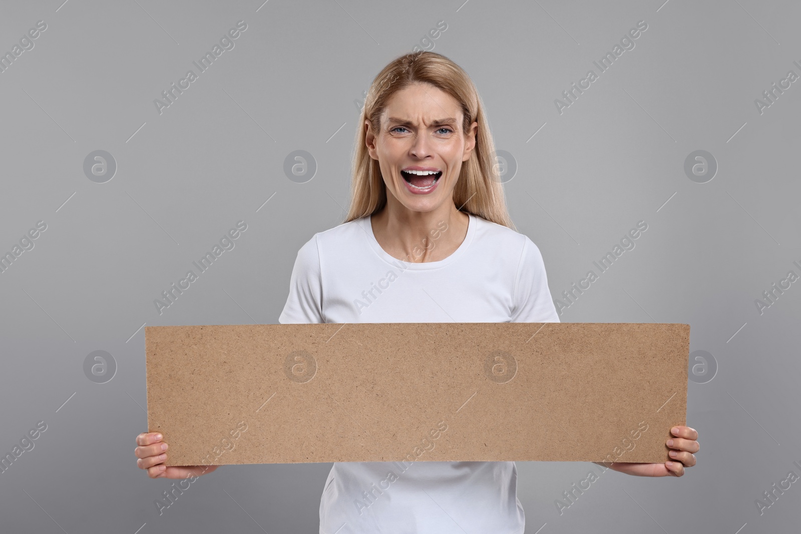 Photo of Angry woman holding blank cardboard banner on grey background, space for text