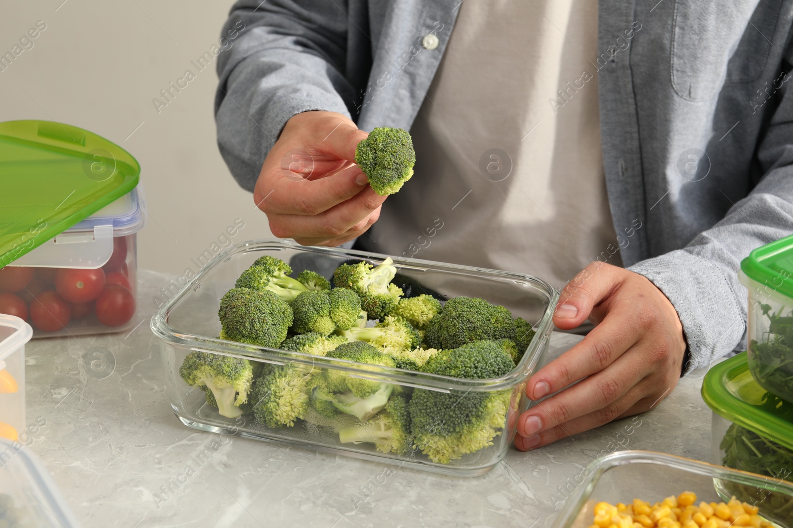 Photo of Man putting fresh broccoli into glass container at light grey table, closeup. Food storage